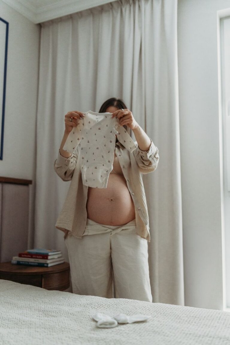 Mama w ciazy trzyma ubranko niemowlece podczas domowej sesji ciazowej w sypialni w Warszawie - Magda Zaborowska Fotografia