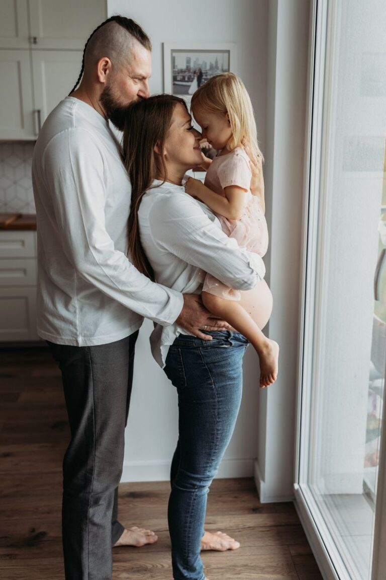 Rodzina podczas domowej sesji ciazowej w Warszawie - tata i mama w ciazy przytulaja coreczke przy oknie - Magda Zaborowska Fotografia