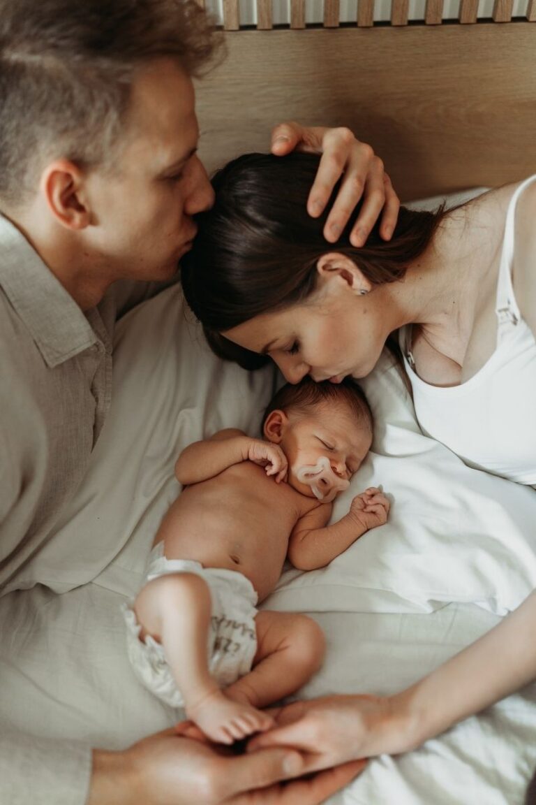 Mama i tata caluja spiacy noworodek lezacy na lozku podczas domowej sesji noworodkowej w Warszawie - Magda Zaborowska Fotografia
